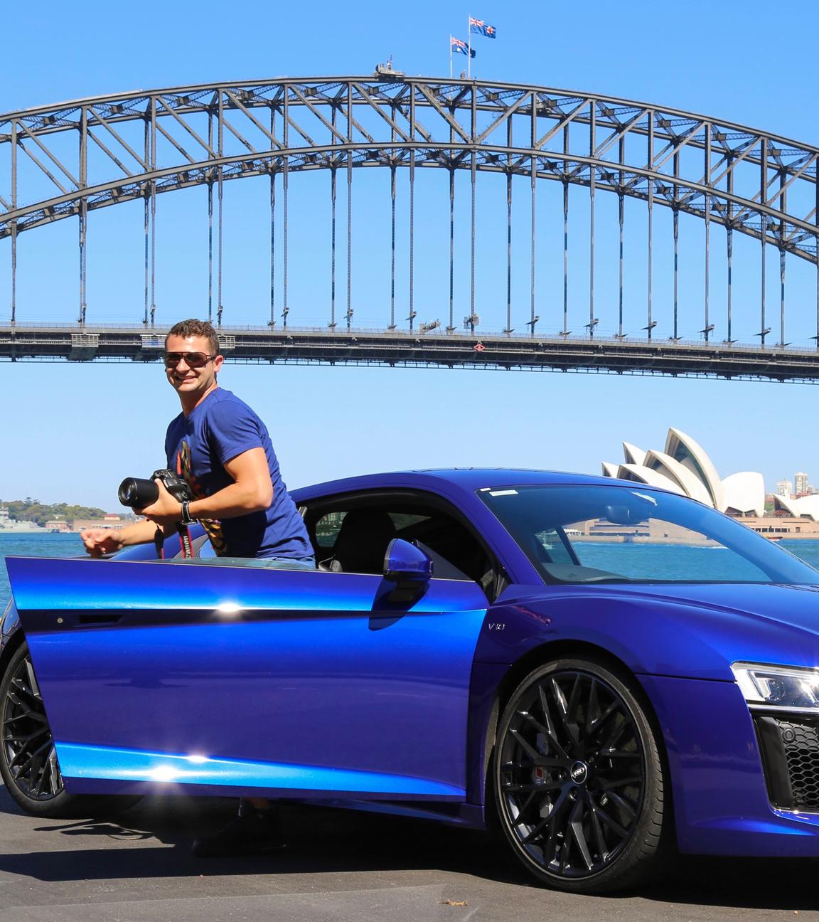 Anthony Koletti steigt aus seinem blauen Sportwagen aus. Im Hintergrund steht die Oper von Sydney, davor führt eine Brücke über ein Gewässer.