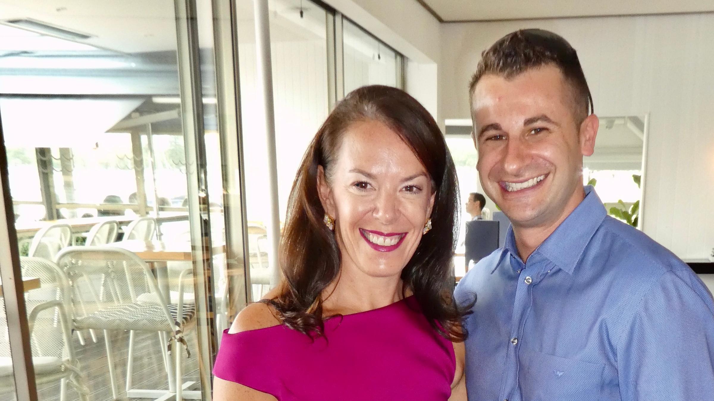 Eine brünette Frau in einem weinroten Kleid mit kurzen Ärmeln und ein dunkelhaariger Mann in blauem Hemd und schwarzer Hose posieren eng aneinanderstehend für ein Foto. 