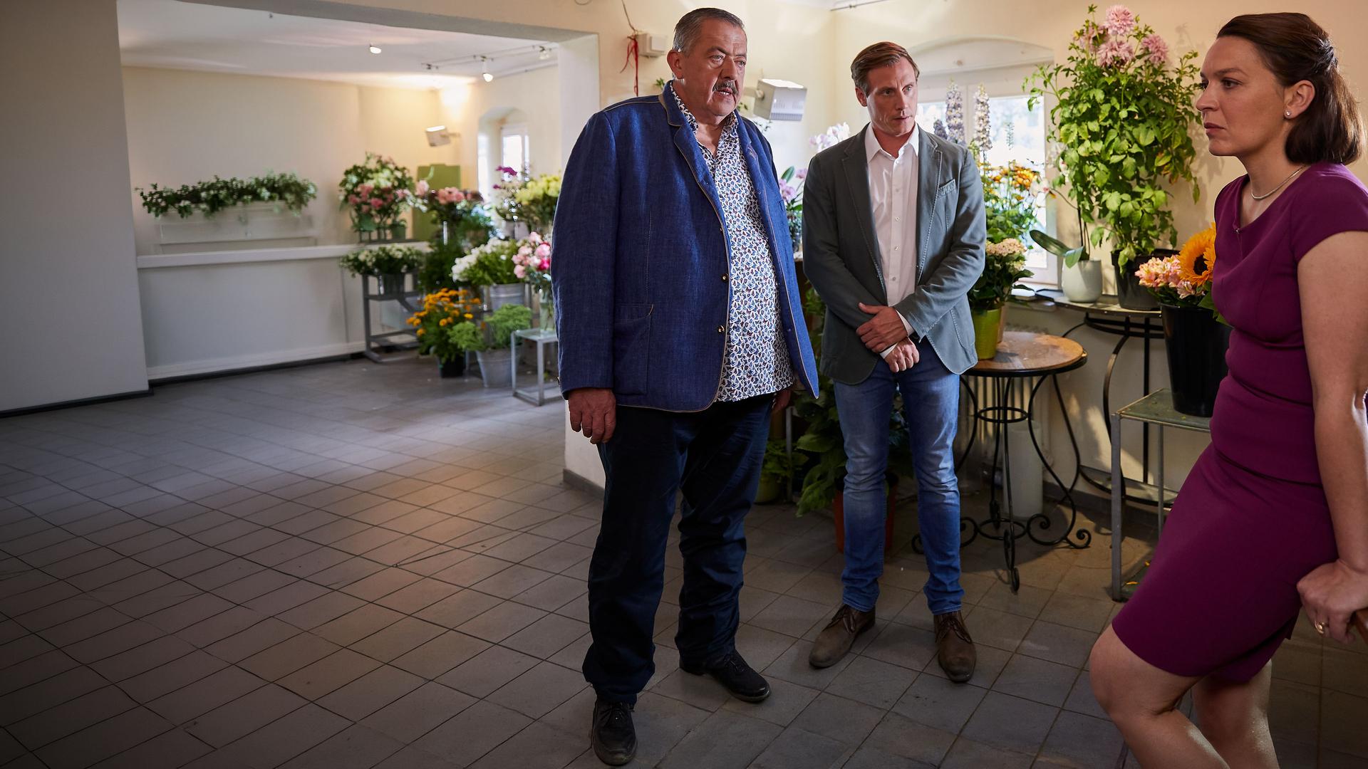 "Die Rosenheim-Cops - Eine verhängnisvolle Spende": Die Kommissare Hofer (Joseph Hannesschläger) und Bach (Patrick Kalupa) stehen in einem Blumenladen nebeneinander und sehen Diana Steinmann (Barbara Romaner) an, die ihnen mit versteinertem Gesicht gegenübersteht und sich auf eine hölzerne Arbeitsplatte stützt.