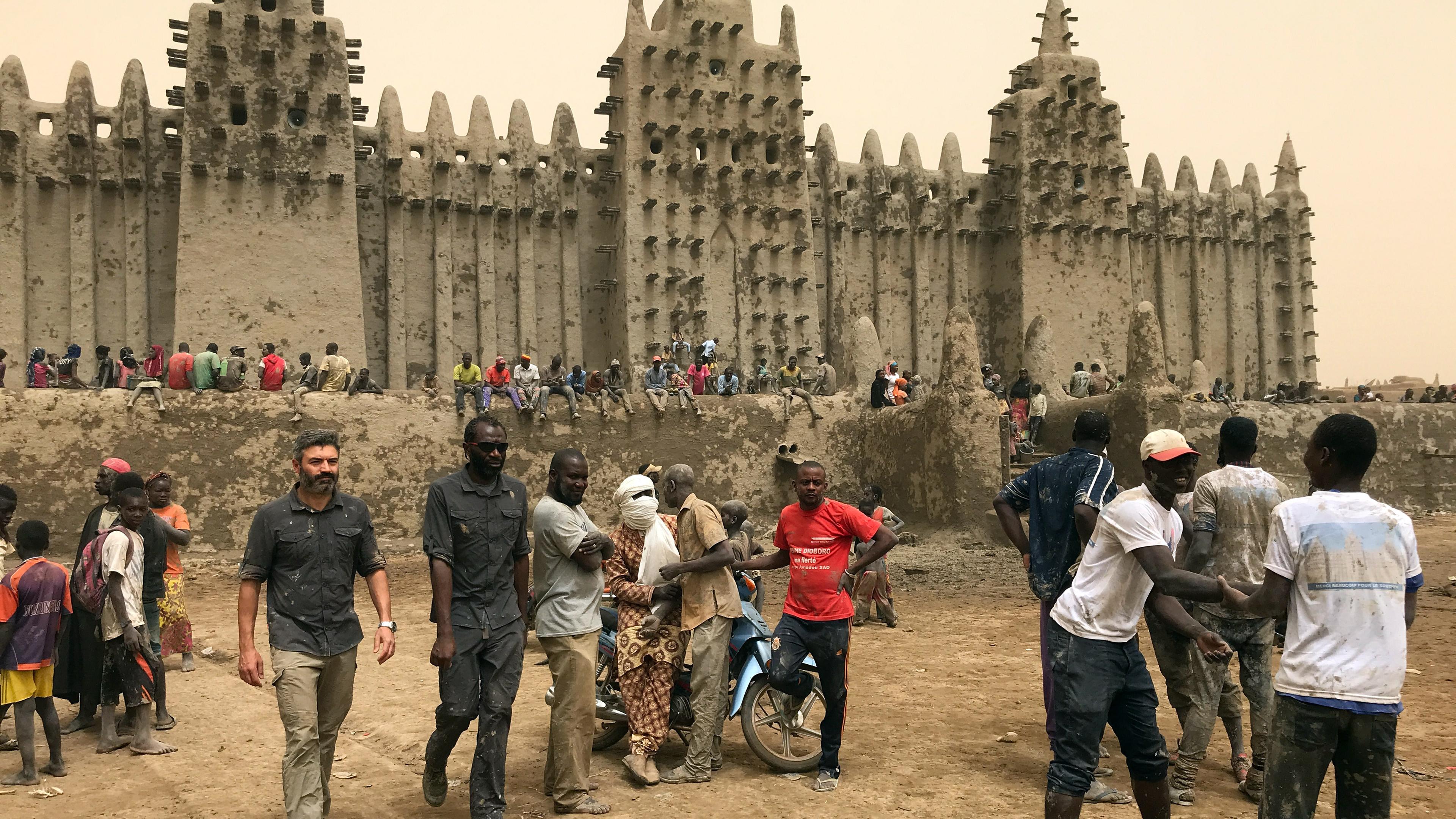 Die imposante Lehmfassade der Moschee von Djenné. Auf der Mauer davor sitzen dichtgedrängt Menschen. Im Vordergrund stehen mehrere Männer.