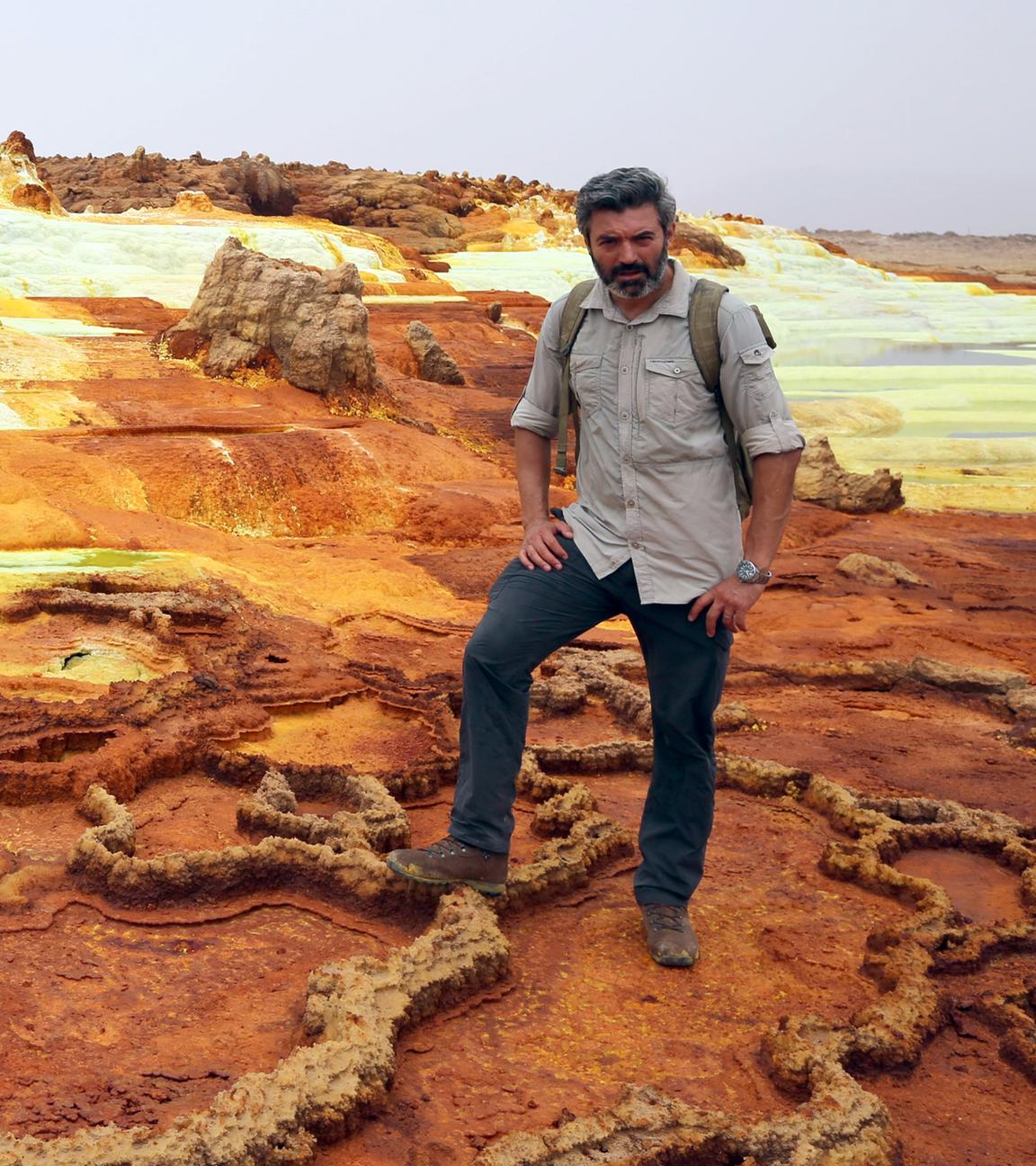 Reporter Reza Pakravan steht in einem Geothermalgebiet, das von krustigen Ablagerungen in Rot-, Braun- und Gelbtönen durchzogen ist, an der Seite liegt ein Schwefelsee.