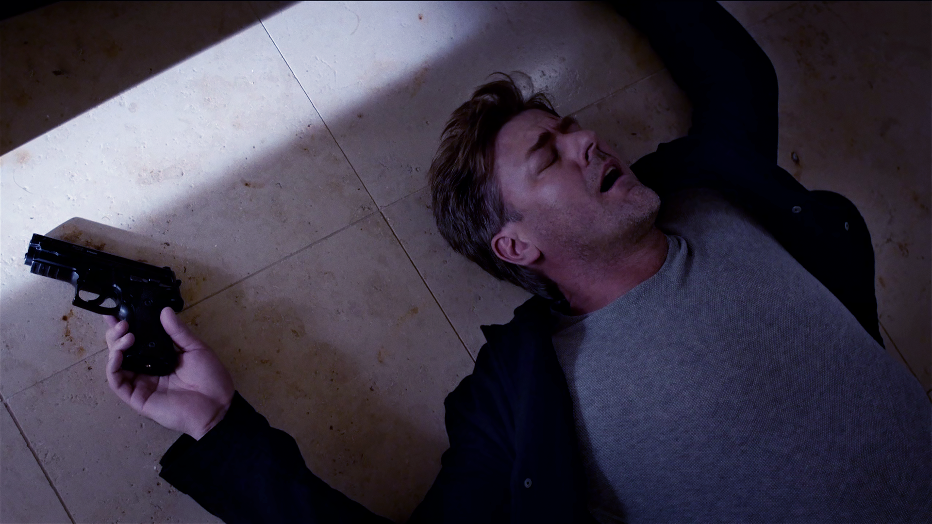 Harald Neuhauser (Marcus Mittermeier) liegt rücklings auf Steinfliesen, in der rechten Hand seine Waffe. 