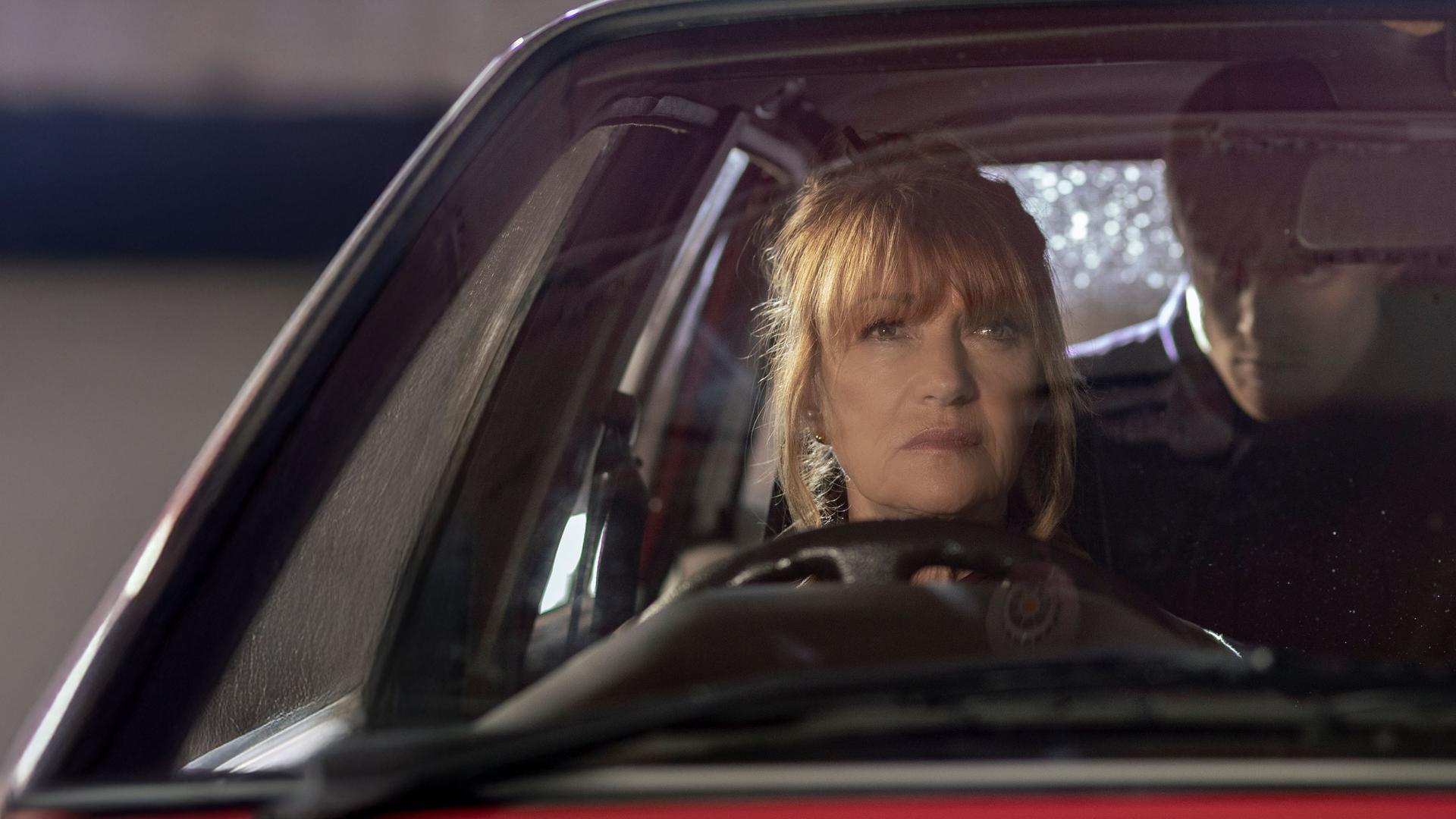 "Harry Wild - Mörderjagd in Dublin: Einer wird gewinnen": Harry Wild (Jane Seymour) hinter dem Steuer eines Autos sitzend und in den Rückspiegel blickend. Hinter ihr, auf der Rückbank des Autos, sitzt ein maskierter, dunkel gekleideter  Mann.