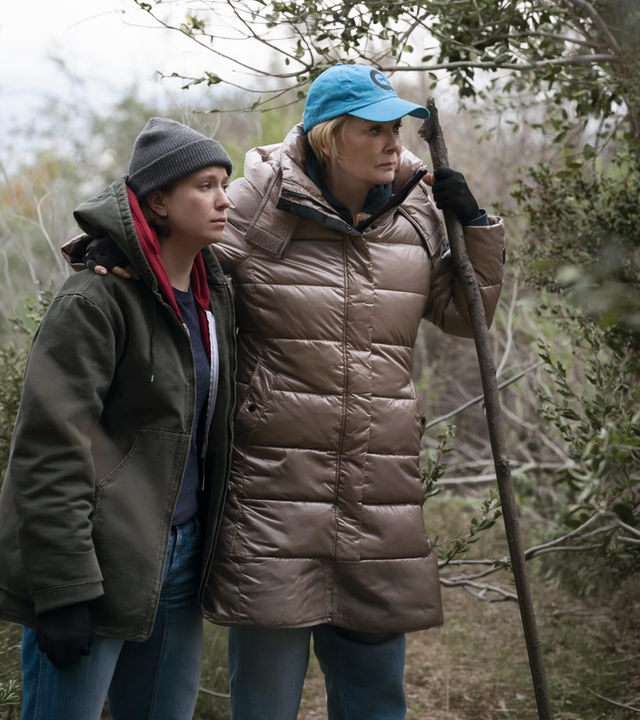 "Hacks - Eines Tages ist jetzt": Deborah und Ava stehen in Outdoorkleidung nebeneinander. Deborah hält in ihrer Hand einen großen Stock, auf den sie sich zu stützt. Um sie herum stehen Bäume und Sträucher.
