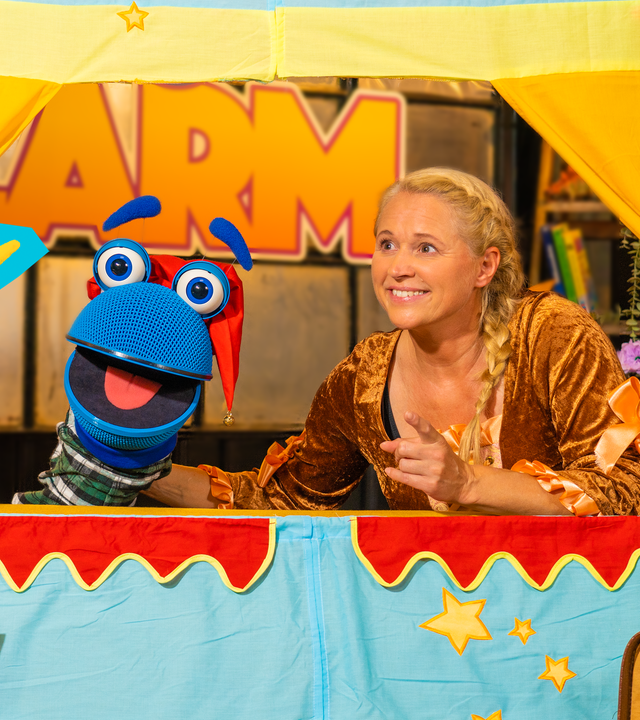 Singa und das SingDing schauen aus einer bunten Bühne für Puppenspiele hervor. Das SingDing hat eine rote Kasper-Mütze auf dem Kopf und Singa trägt ein orangebraunes Samtkleid wie eine Märchenprinzessin. Beide schauen freudig zu herrH, der vor der Bühne steht. Er trägt gelbe Putzhandschuhe und hält eine Putzbürste in der Hand. Er lacht und hat die Arme leicht ausgebreitet. Auf seinem violetten Pullover ist ein großes, türkisfarbenes „H“ abgebildet.