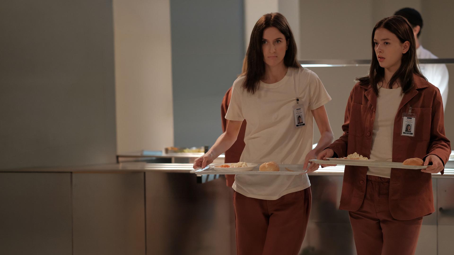 "Fallen - Eingewöhnung": Luce (Jessica Alexander) und Arriane (Josefine Koenig) stehen in der Kantine und tragen jeweils ein Tablett mit Essen. Beide blicken ernst, während hinter ihnen ein Mitarbeiter (Komparse) an der Essensausgabe arbeitet.