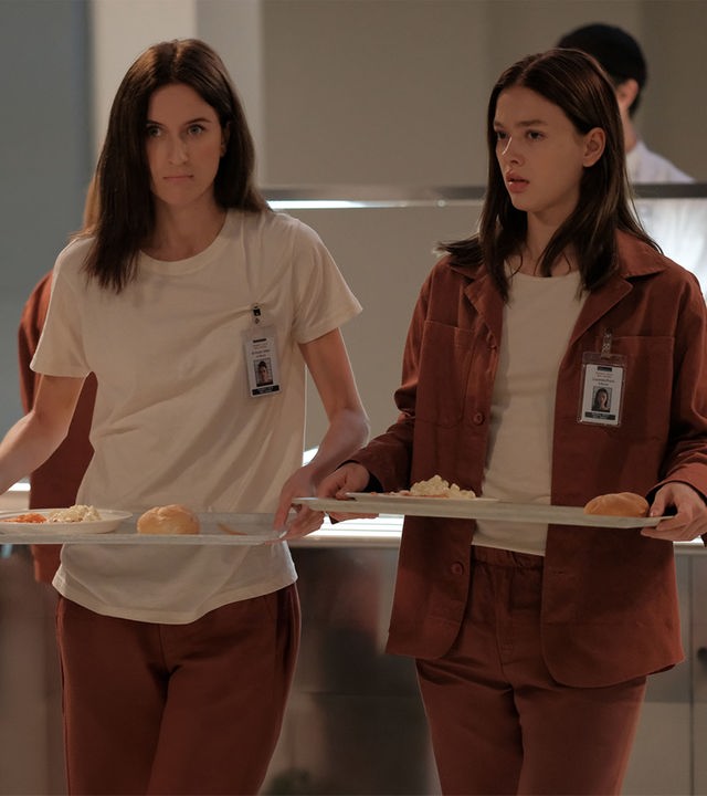 "Fallen - Eingewöhnung": Luce (Jessica Alexander) und Arriane (Josefine Koenig) stehen in der Kantine und tragen jeweils ein Tablett mit Essen. Beide blicken ernst, während hinter ihnen ein Mitarbeiter (Komparse) an der Essensausgabe arbeitet.
