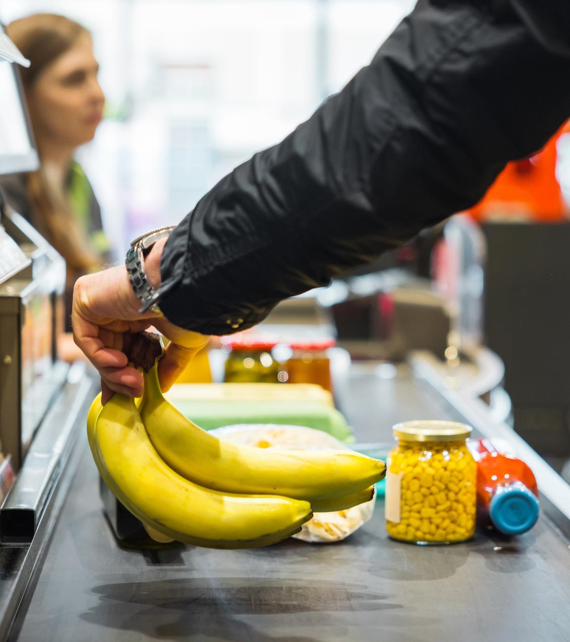 Person legt Bananen zusammen mit anderen Artikeln auf das Förderband an der Supermarktkasse, während im Hintergrund eine Kassiererin zu sehen ist.
