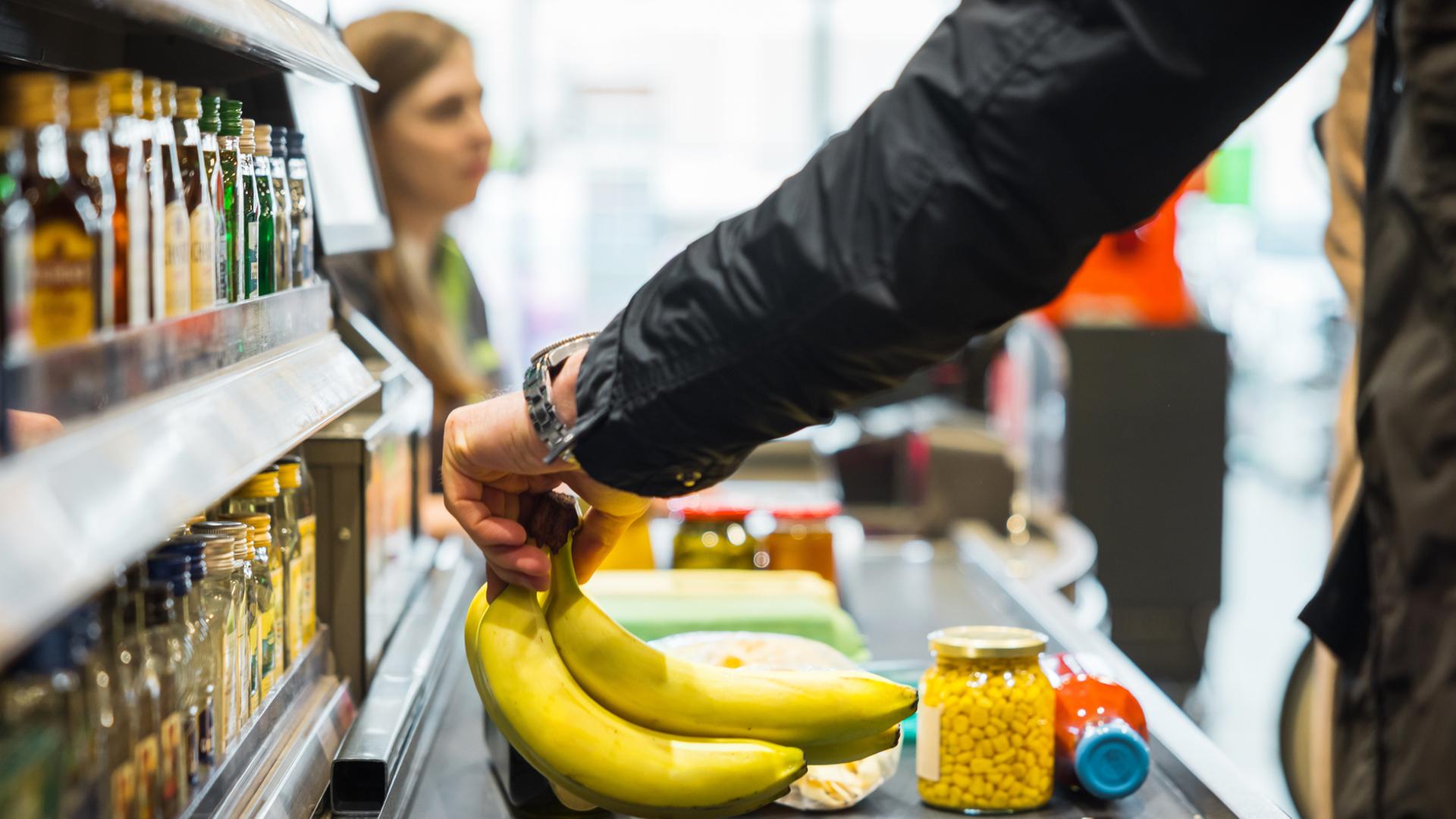 Person legt Bananen zusammen mit anderen Artikeln auf das Förderband an der Supermarktkasse, während im Hintergrund eine Kassiererin zu sehen ist.