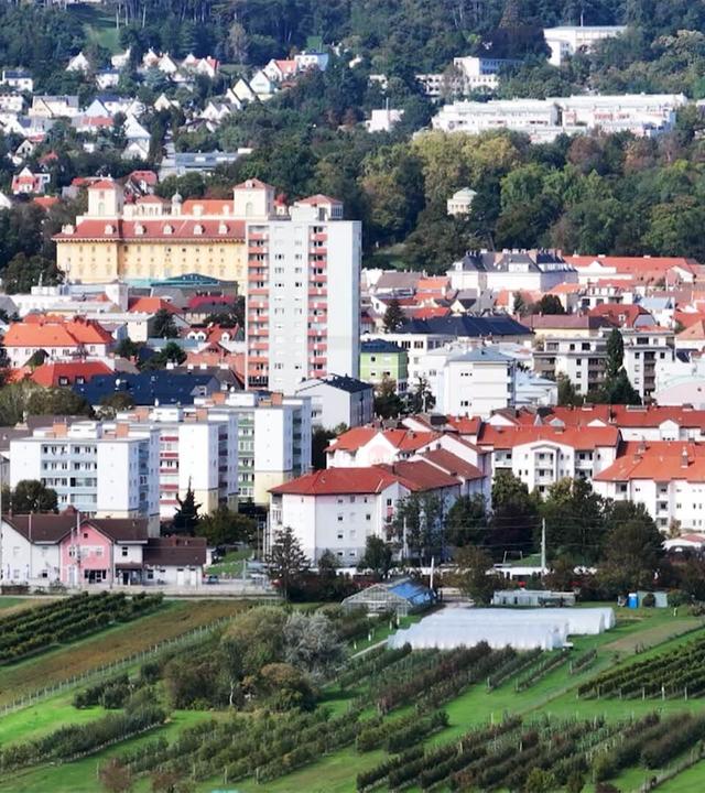 Das Bild zeigt eine Panoramaansicht von Eisenstadt, der Landeshauptstadt des Burgenlandes in Österreich. Im Vordergrund sind ausgedehnte grüne Wiesen und Weinberge zu sehen, die in geschwungenen Reihen angeordnet sind.   Im Hintergrund ist die Stadt sichtbar, bestehend aus verschiedenen Gebäuden mit unterschiedlichen Architekturstilen. Viele der Häuser haben rote Dächer, einige sind mehrstöckig, und es gibt auch modernere Wohnhäuser. In zentraler Lage fällt ein großes, gelb gestrichenes Gebäude mit symmetrischen Fenstern auf, das vermutlich das Schloss Esterhazy darstellt.   Auf der rechten Seite des Bildes steht ein Kirchturm, der mit einem roten Dach versehen ist. Die Umgebung ist von grünen Hügeln und Bäumen gesäumt, was das Bild insgesamt harmonisch und malerisch wirken lässt. Der Himmel ist größtenteils klar, was auf einen sonnigen Tag hindeutet.   Die Bildunterschrift weist darauf hin, dass Eisenstadt als "die kleinste Großstadt der Welt" beworben wird und feiert 100 Jahre als Landeshauptstadt.