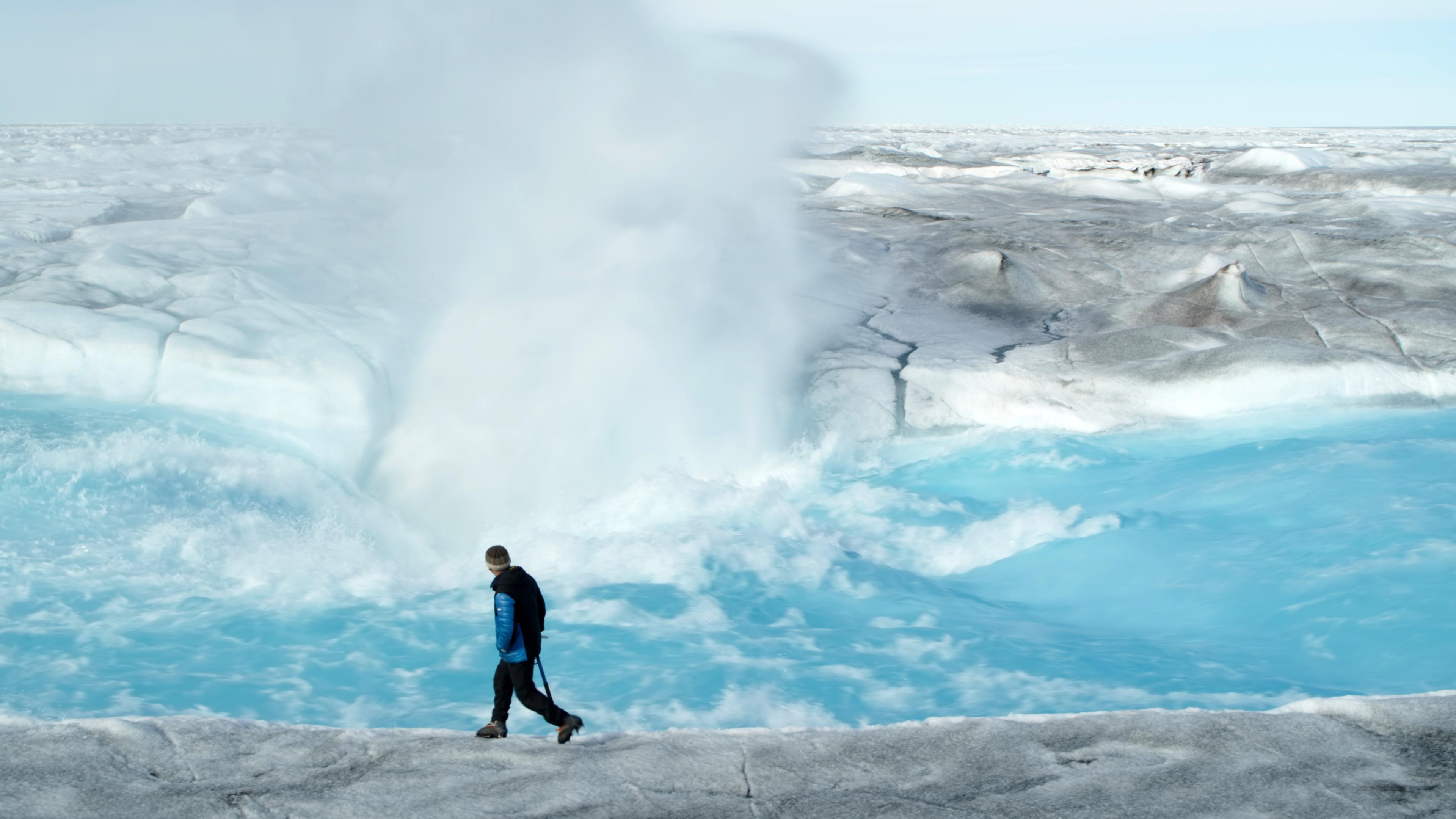 Glaziologe Prof. Alun Hubbard steht vor einem reißenden Schmelzwasserfluss auf dem Eis, während hinter ihm tosend Gischt emporsteigt.
