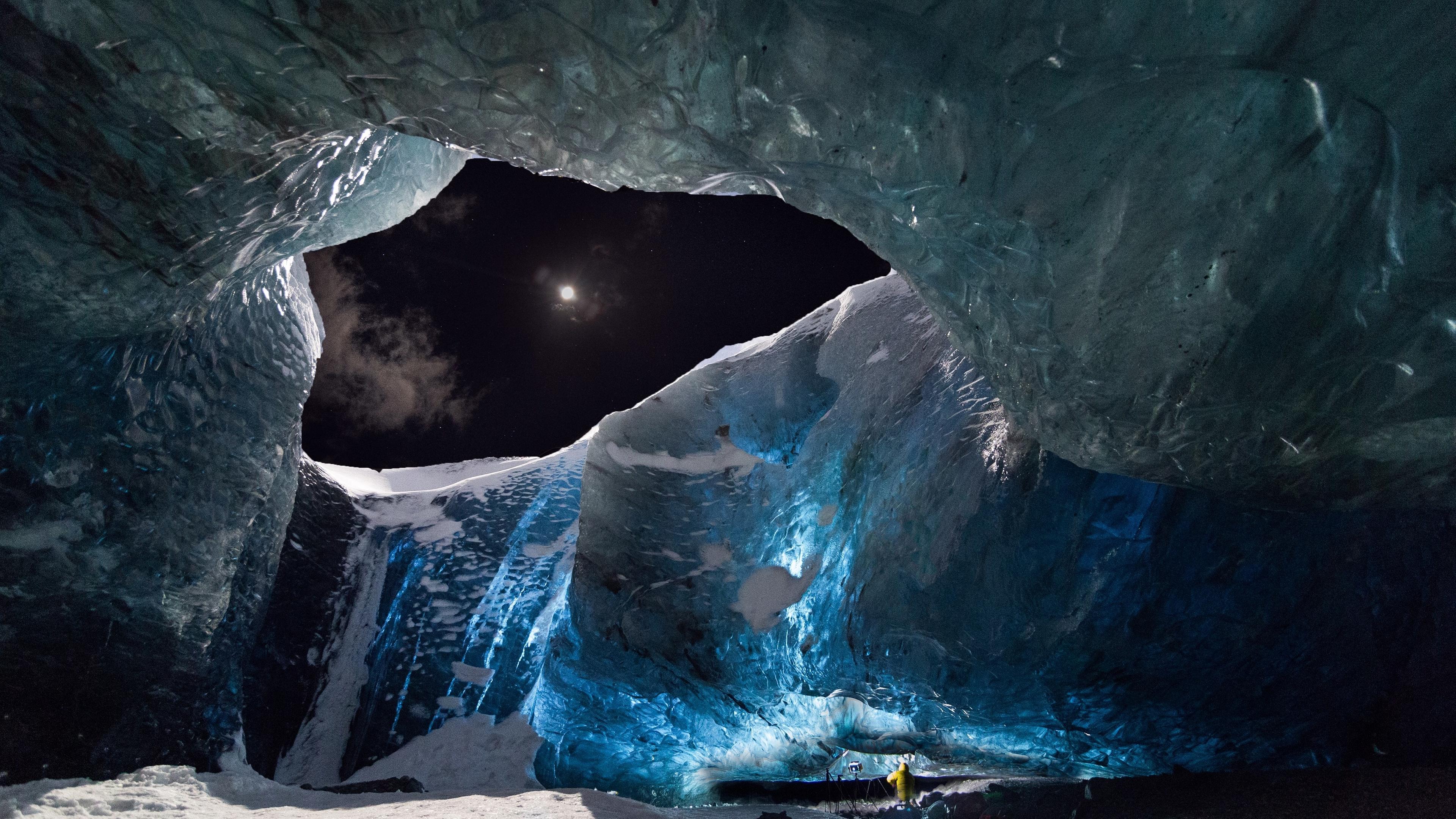 Bei Nacht bereitet sich ein Kamerateam unter einem meterdicken isländischen Gletscher auf die Dreharbeiten vor. Durch eine Öffnung im Gletscher ist am Nachthimmel der Mond zu sehen.