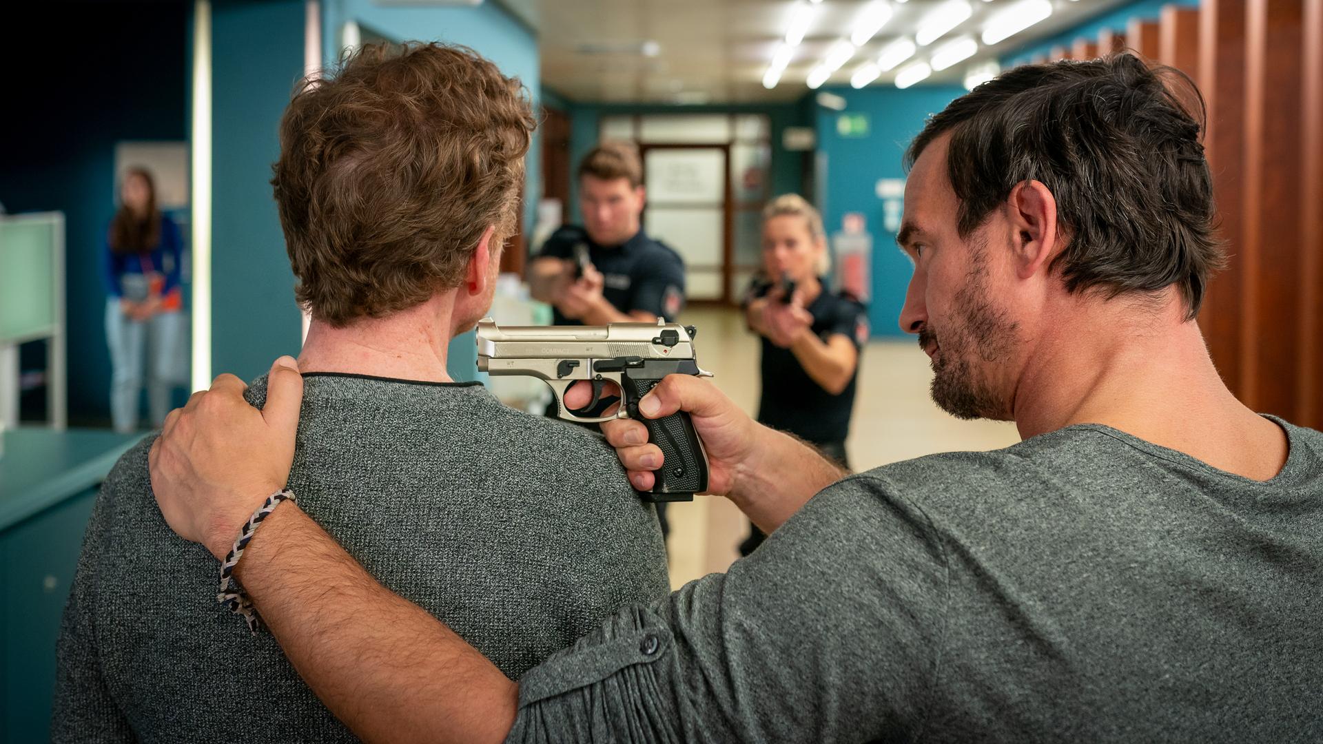 Im Flur des EKH: Matthias Engel (Sönke Möhring) stehet dicht neben Anton Heissner (Mathias Junge). Er hat seine linke Hand auf dessen linken Schulter liegen. In der Rechten hält er eine Pistole, die er auf ihn gerichtet hat. Er blickt ihn von der Seite an. Etwas entfernt vor ihnen stehen der Polizist Nick Brandt (Raúl Richter) und seine Kollegin Franzi Jung (Rhea Harder-Vennewald). Sie stehen nebeneinander. Beide haben ihre Waffe auf beide Männer gerichtet.