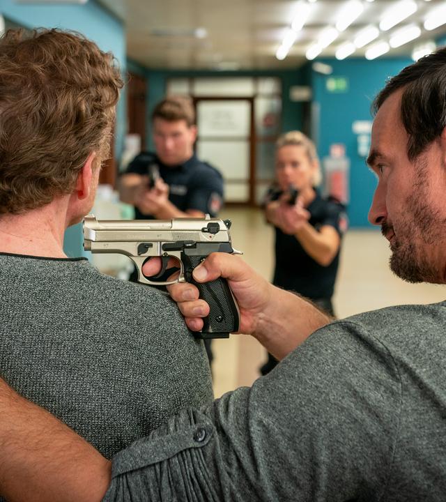 Im Flur des EKH: Matthias Engel (Sönke Möhring) stehet dicht neben Anton Heissner (Mathias Junge). Er hat seine linke Hand auf dessen linken Schulter liegen. In der Rechten hält er eine Pistole, die er auf ihn gerichtet hat. Er blickt ihn von der Seite an. Etwas entfernt vor ihnen stehen der Polizist Nick Brandt (Raúl Richter) und seine Kollegin Franzi Jung (Rhea Harder-Vennewald). Sie stehen nebeneinander. Beide haben ihre Waffe auf beide Männer gerichtet.