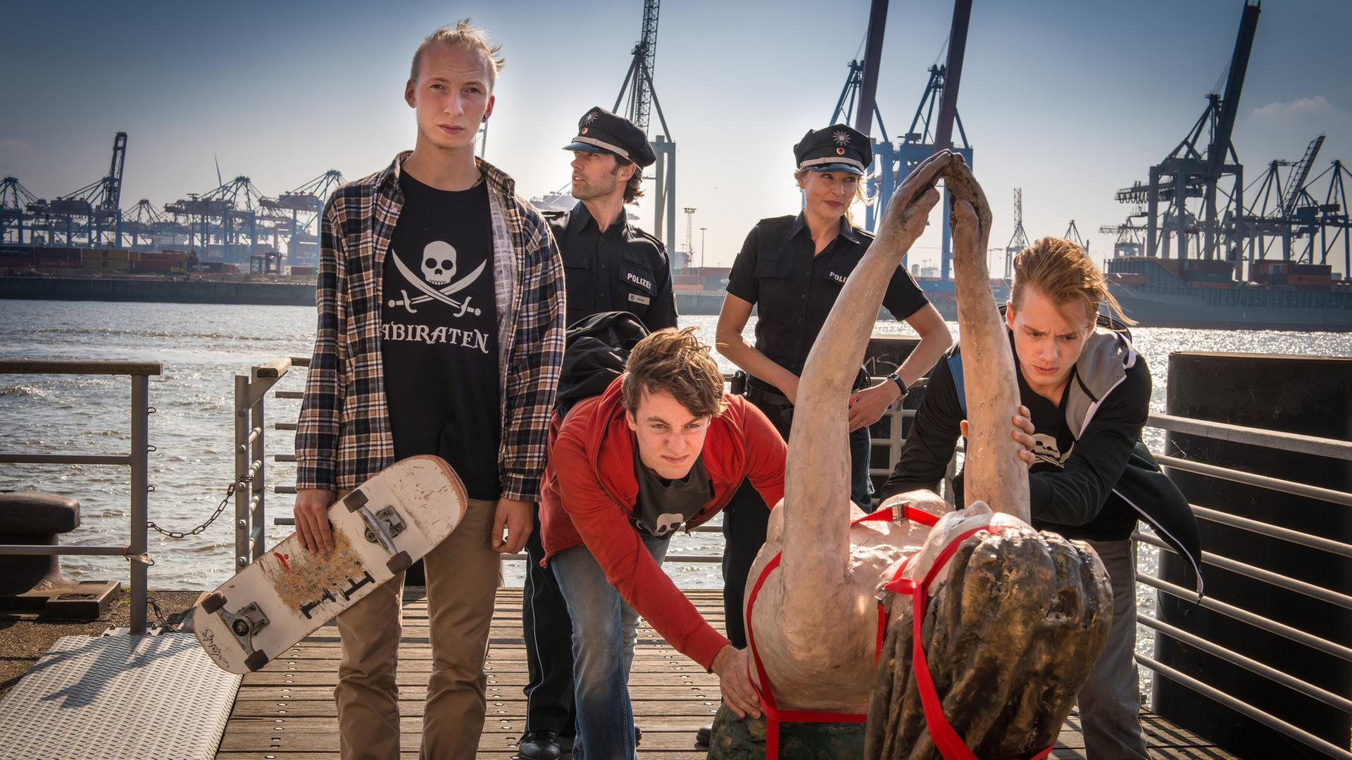 "Notruf Hafenkante - Elbnixe": Die Polizeibeamten Mattes Seeler (Matthias Schloo) und Melanie Hansen (Sanna Englund) schauen zu, wie Paul Heffner (Cecil von Renner) und seine zwei Freunde eine Galionsfigur über ein Schiff schieben. Im Hintergrund der Hamburger Hafen.