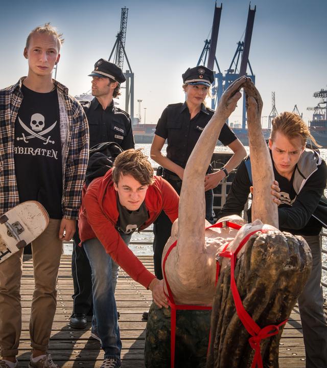 "Notruf Hafenkante - Elbnixe": Die Polizeibeamten Mattes Seeler (Matthias Schloo) und Melanie Hansen (Sanna Englund) schauen zu, wie Paul Heffner (Cecil von Renner) und seine zwei Freunde eine Galionsfigur über ein Schiff schieben. Im Hintergrund der Hamburger Hafen.