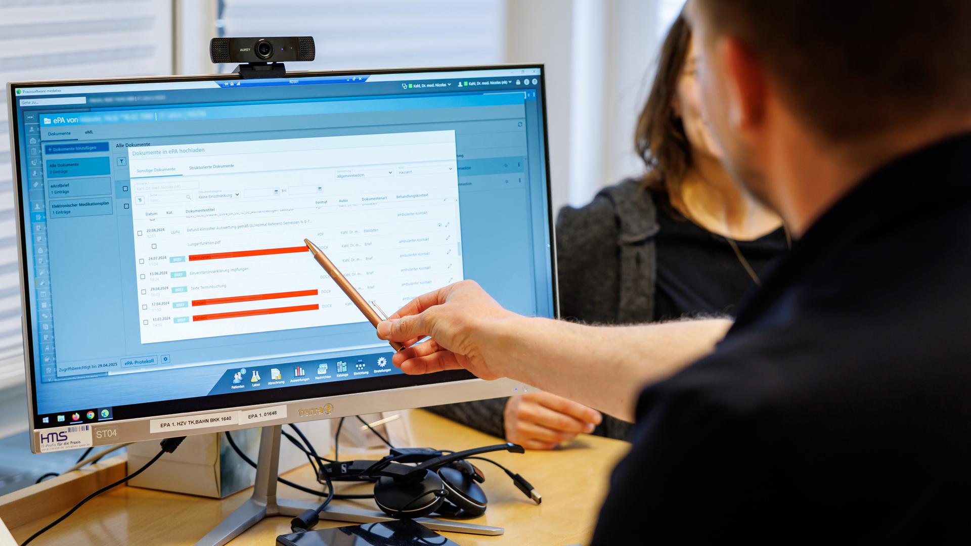 Hausarzt (von hinten zu sehen) lädt Dokumente in eine elektronische Patientenakte "ePA", sitzt vor Computer und zeigt mit Stift darauf
