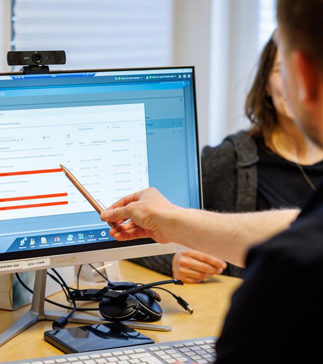 Hausarzt (von hinten zu sehen) lädt Dokumente in eine elektronische Patientenakte "ePA", sitzt vor Computer und zeigt mit Stift darauf
