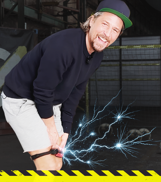 Tommy hat einen Elektroschocker um sein Knie gebunden und lächelt in die Kamera. Im Hintergrund steht ein Fußballtor.