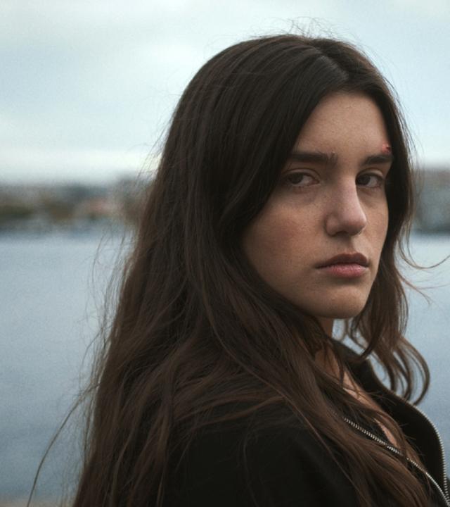 Hazal (Melia Kara) steht an einem See und blickt mit einem verunsiocherten Gesichtsausdruck in die Kamera.