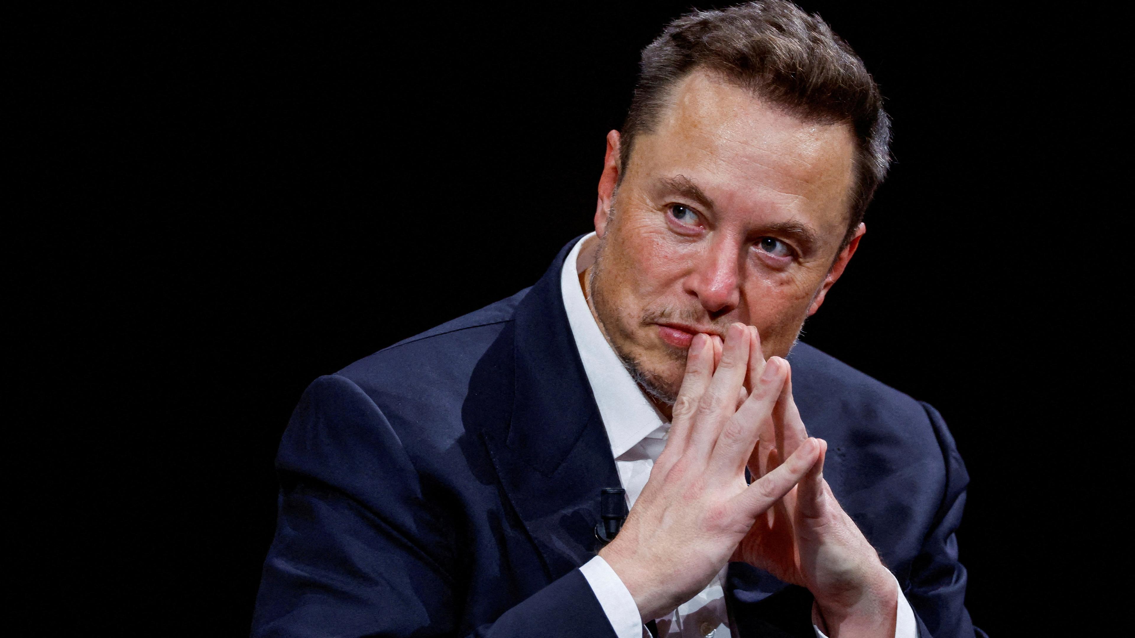 Elon Musk hinter schwarzem Hintergrund mit zusammengefalteten Händen.