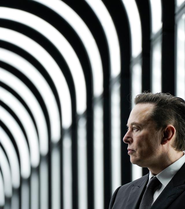 Ein Mann mittleren Alters im grauen Anzug steht vor einem schwarz-weiß-gestreiften Hintergrund.