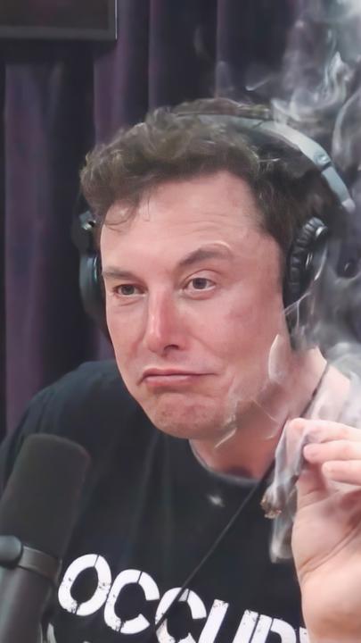 Elon Musk, mit schwarzem T-Shirt mit weißer Aufschrift, sitzt während eines Interviews im Jahr 2018 vor einem Mikrophon und hält in der linken Hand einen Joint, der qualmt.