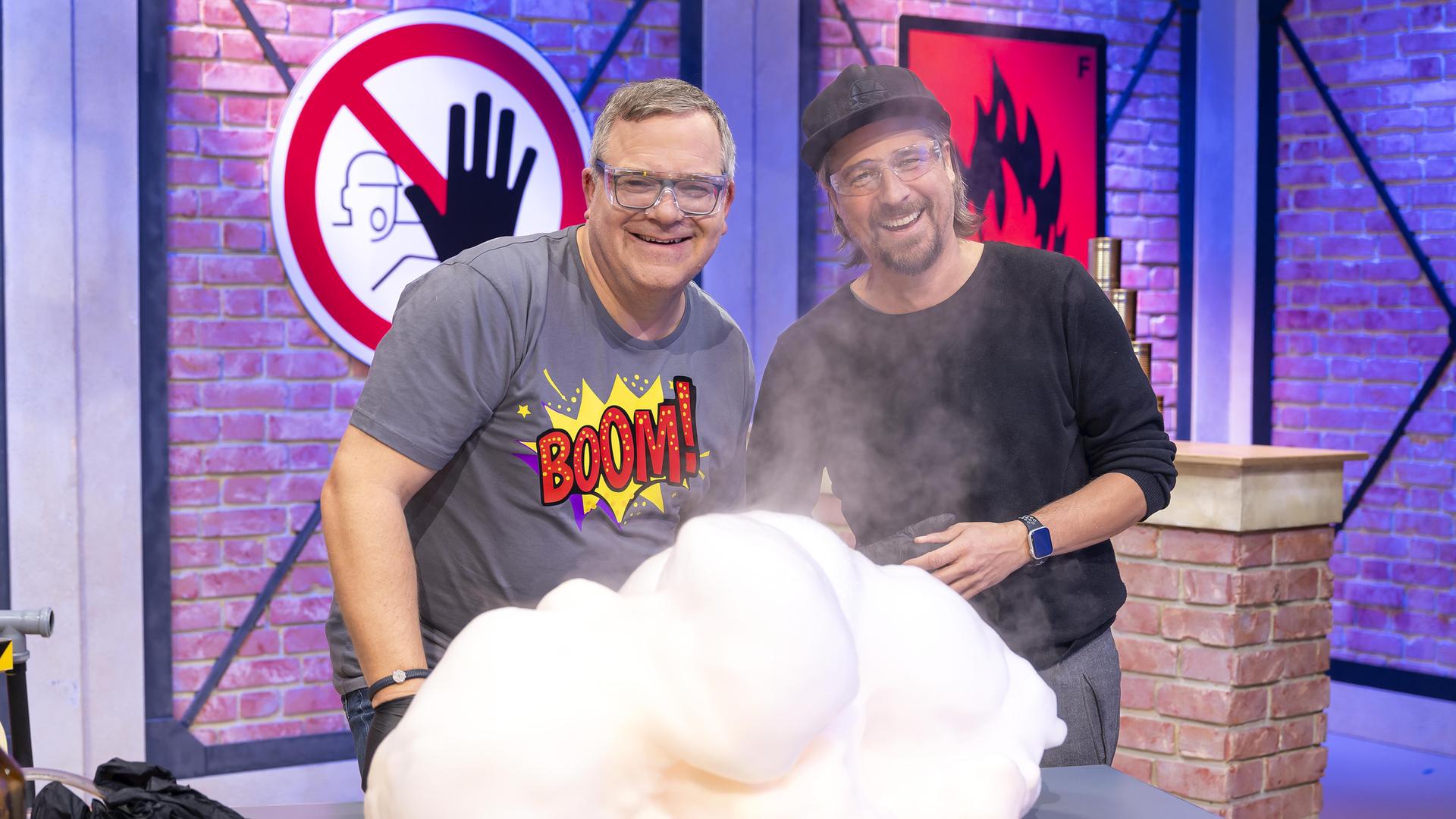 Elton und Gast Tommy Scheel stehen vor einem Labortisch und tragen Sicherheitsbrillen. Vor ihnen raucht ein Berg aus Schaum. Beide lachen.