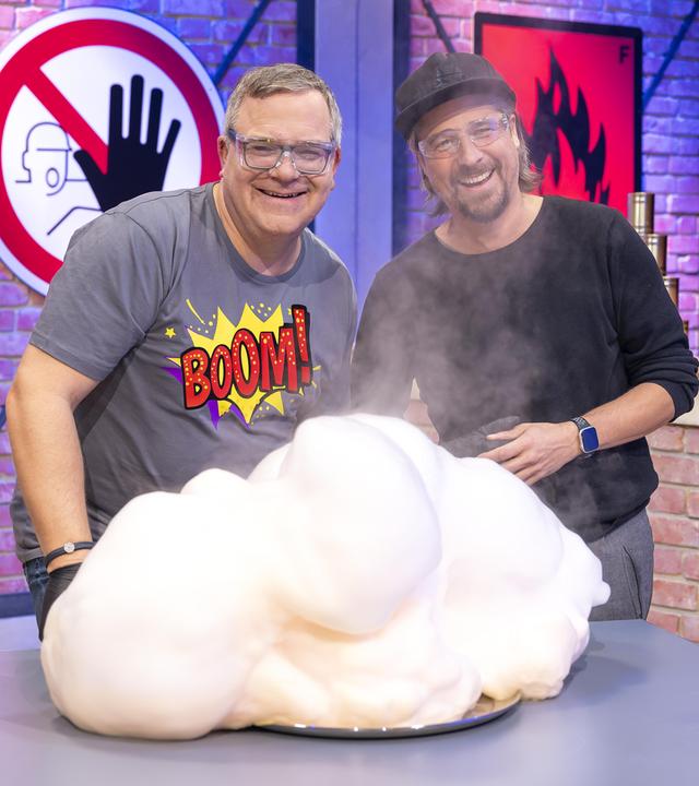 Elton und Gast Tommy Scheel stehen vor einem Labortisch und tragen Sicherheitsbrillen. Vor ihnen raucht ein Berg aus Schaum. Beide lachen.