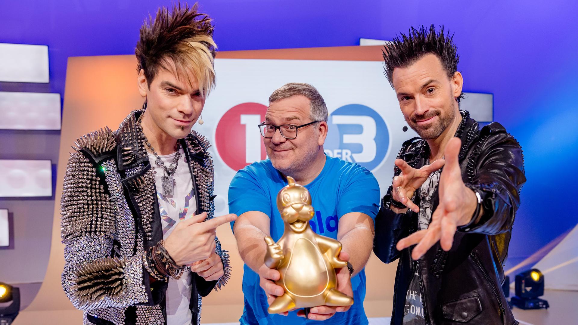 Die Ehrlich Brothers Chris und Andreas stehen neben Elton und machen Zaubergesten. Elton hält stolz eine goldene Piet-Flosse-Figur in die Kamera.