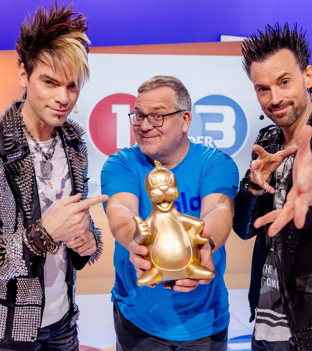 Die Ehrlich Brothers Chris und Andreas stehen neben Elton und machen Zaubergesten. Elton hält stolz eine goldene Piet-Flosse-Figur in die Kamera.