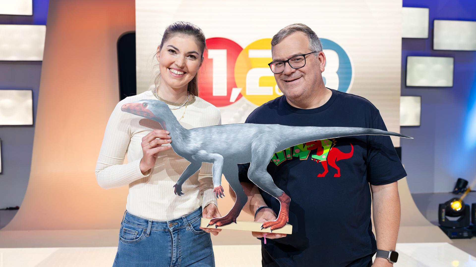 "1, 2 oder 3 - Elton und die Welt der Dinosaurier": Biologin Kathrin Helsper steht mit Elton vor dem "1, 2 oder 3"-Logo im Studio. Zwischen sich halten sie das Modell eines Dinosauriers.