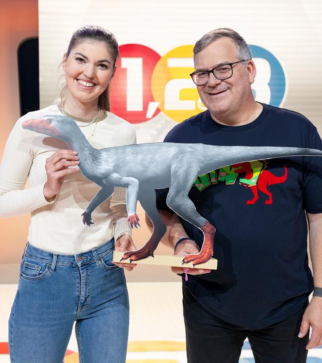 "1, 2 oder 3 - Elton und die Welt der Dinosaurier": Biologin Kathrin Helsper steht mit Elton vor dem "1, 2 oder 3"-Logo im Studio. Zwischen sich halten sie das Modell eines Dinosauriers.