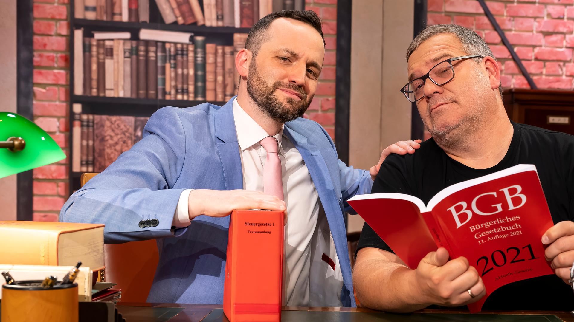 Herr Anwalt" Tim Hendrik Walter und Elton sitzen hinter einem großen Schreibtisch. Beide schauen ernst, aber sympathisch in die Kamera. Auf dem Tisch stehen mehrere Gesetzbücher und eine kleine Statue der römischen Göttin Justitia. Elton hält ein großes Gesetzbuch in den Händen.