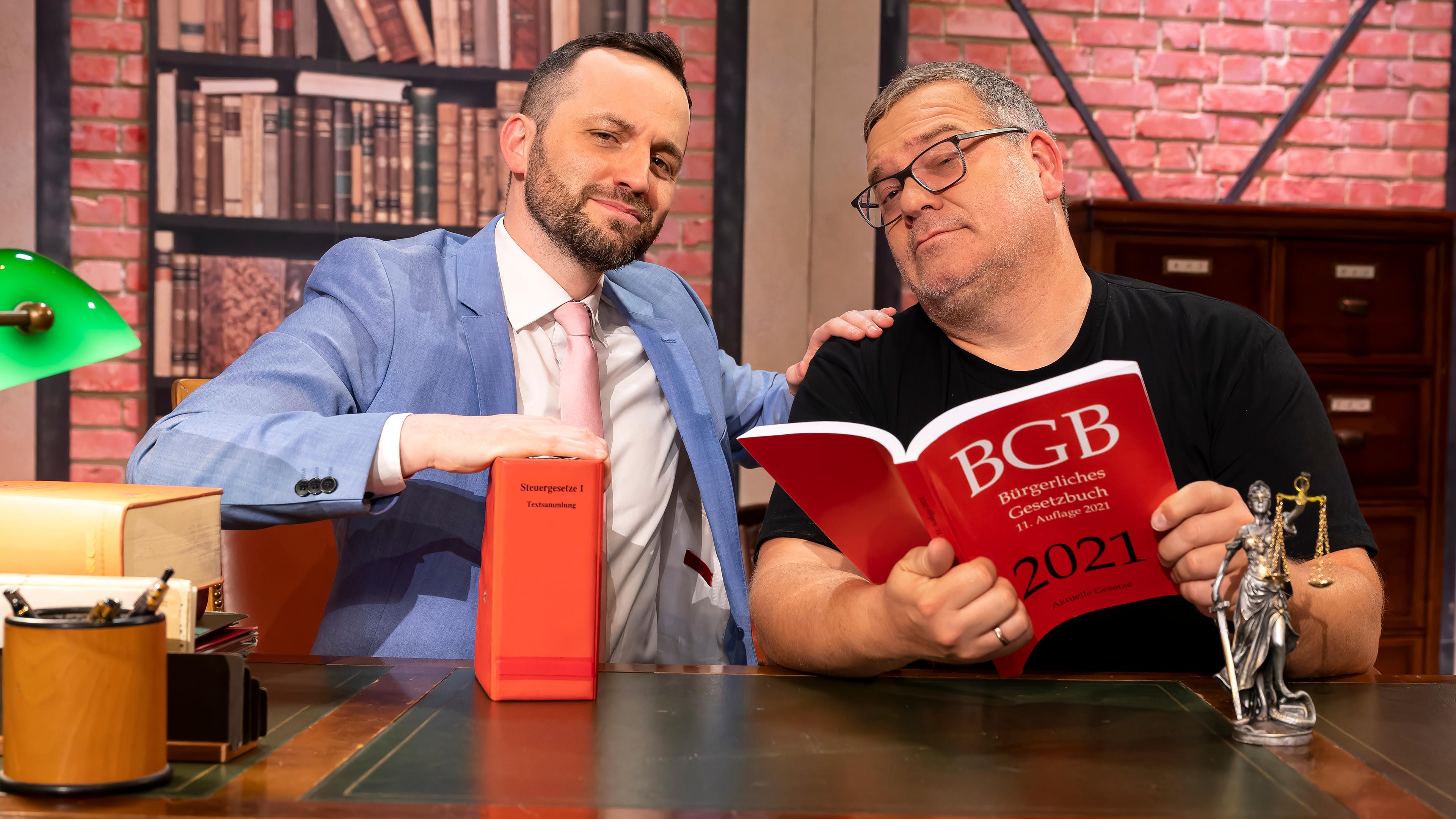 Herr Anwalt" Tim Hendrik Walter und Elton sitzen hinter einem großen Schreibtisch. Beide schauen ernst, aber sympathisch in die Kamera. Auf dem Tisch stehen mehrere Gesetzbücher und eine kleine Statue der römischen Göttin Justitia. Elton hält ein großes Gesetzbuch in den Händen.