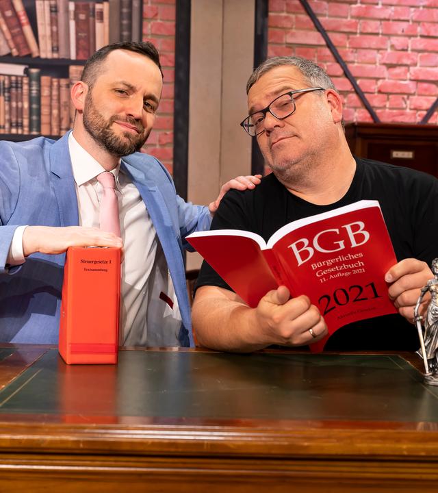 Herr Anwalt" Tim Hendrik Walter und Elton sitzen hinter einem großen Schreibtisch. Beide schauen ernst, aber sympathisch in die Kamera. Auf dem Tisch stehen mehrere Gesetzbücher und eine kleine Statue der römischen Göttin Justitia. Elton hält ein großes Gesetzbuch in den Händen.