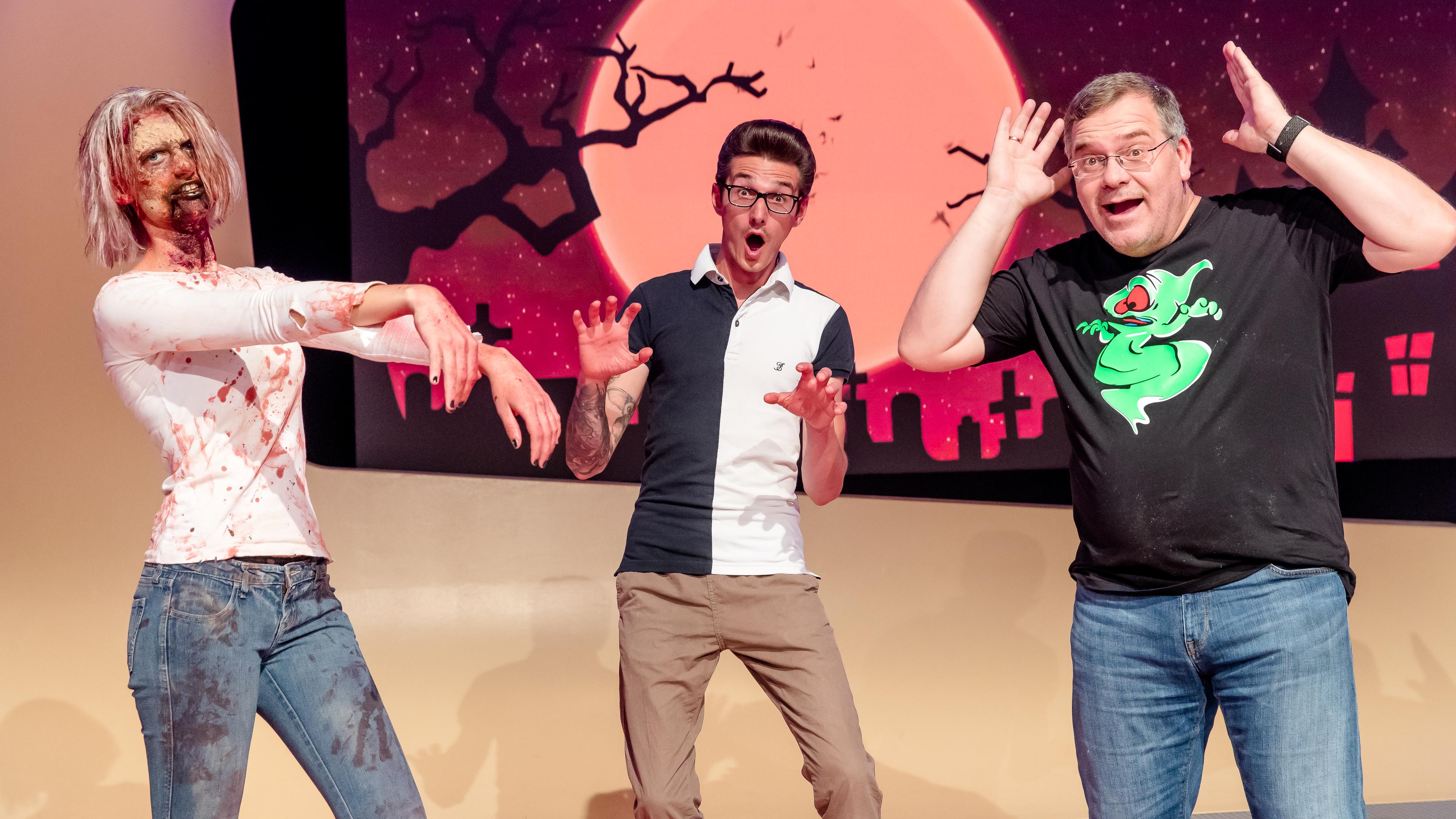 Eine als Zombie geschminkte Frau läuft ins Bild, während Maskenbildner Tim Scheidig verschreckt schaut und Moderator Elton in die Kamera lacht.