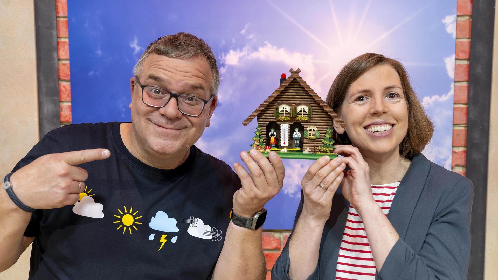 Wetter-Expertin Verena Leyendecker (r.) hält zusammen mit Elton (l.) ein Wetterhäuschen in die Höhe. Beide lachen.