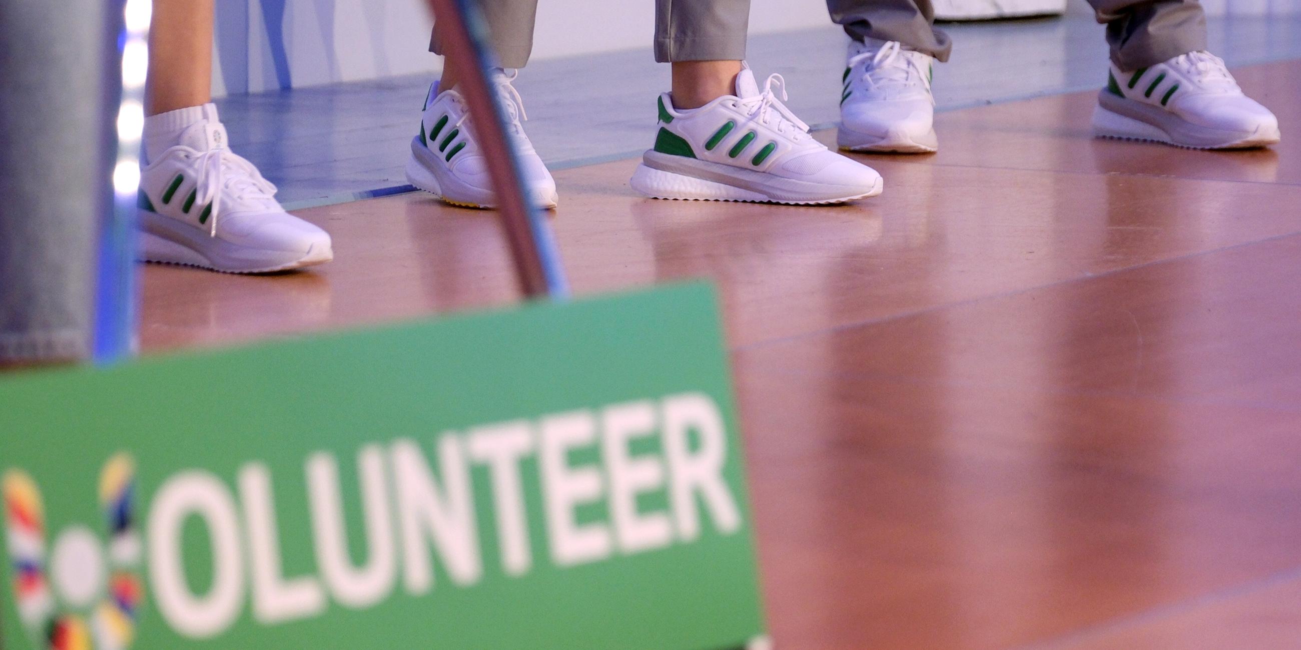 Im Vordergrund ein Schild mit der Aufschrift "Volunteer". Im Hintergrund Beine der ehrenamtlichen Helfer.