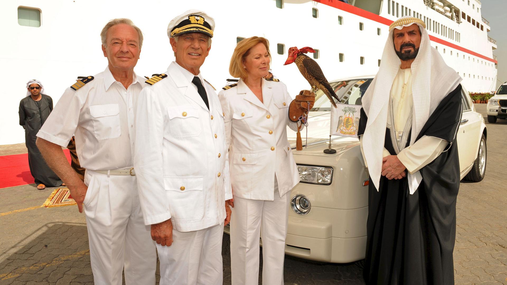 "Das Traumschiff: Emirate": Beatrice (Heide Keller), Dr. Schröder (Horst Naumann) und Kapitän Paulsen (Siegfried Rauch) stehen vor der MS Deutschland. Neben ihnen steht Scheich Youssof (Pierre Semmler), im Hintergrund sein edles Auto. Beatrice hält einen Falken auf der Hand.