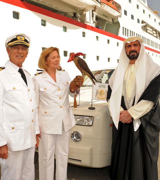 "Das Traumschiff: Emirate": Beatrice (Heide Keller), Dr. Schröder (Horst Naumann) und Kapitän Paulsen (Siegfried Rauch) stehen vor der MS Deutschland. Neben ihnen steht Scheich Youssof (Pierre Semmler), im Hintergrund sein edles Auto. Beatrice hält einen Falken auf der Hand.