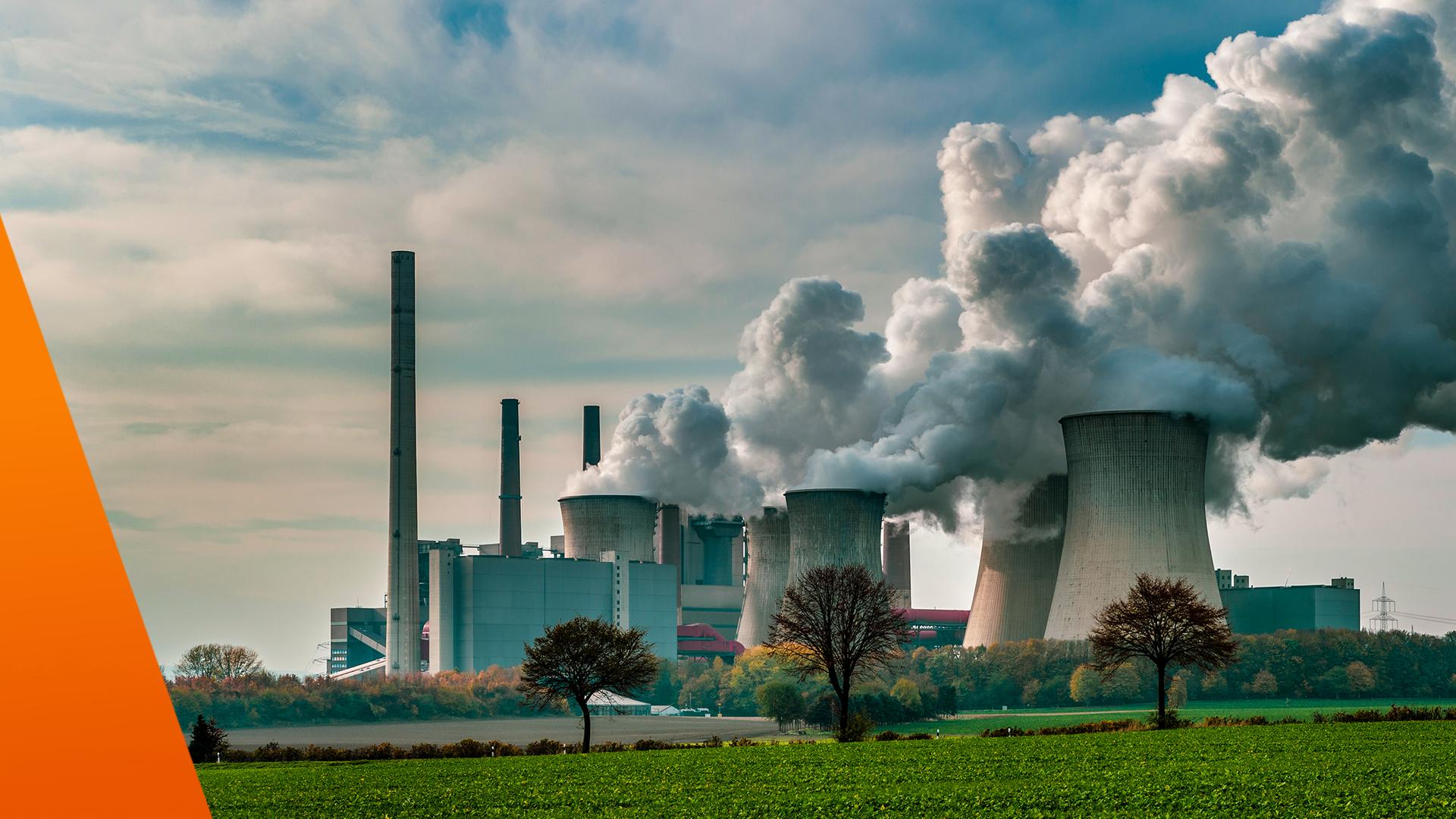 Zu sehen ist ein großes Kraftwerk mit mehreren Kühltürmen und Schornsteinen, aus denen dichte weiße Dampfwolken aufsteigen. Im Vordergrund liegen Felder und vereinzelte Bäume, darüber ein leicht bewölkter Himmel. Das Bild steht sinnbildlich für industrielle CO₂-Emissionen und die Herausforderungen des Emissionshandels.