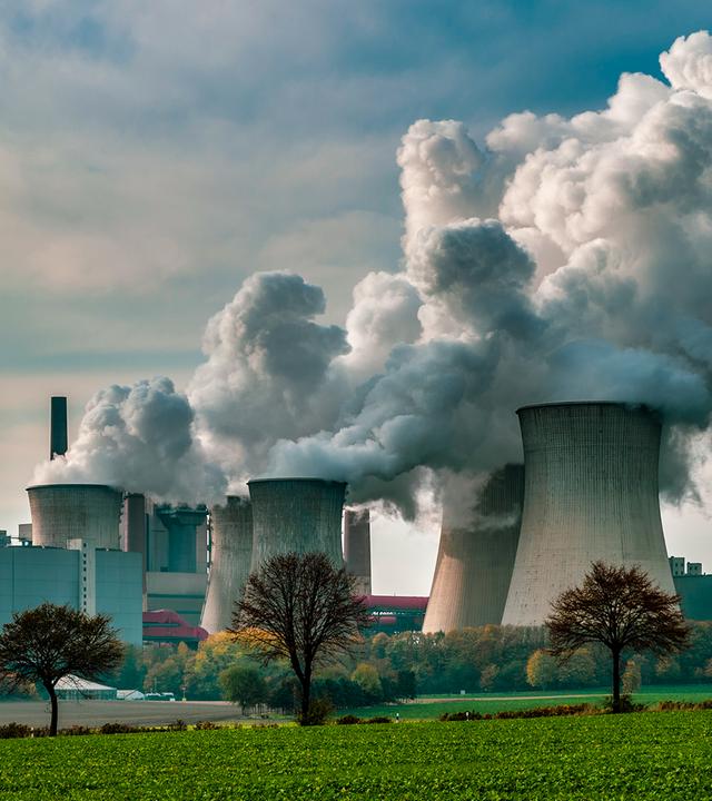 Zu sehen ist ein großes Kraftwerk mit mehreren Kühltürmen und Schornsteinen, aus denen dichte weiße Dampfwolken aufsteigen. Im Vordergrund liegen Felder und vereinzelte Bäume, darüber ein leicht bewölkter Himmel. Das Bild steht sinnbildlich für industrielle CO₂-Emissionen und die Herausforderungen des Emissionshandels.