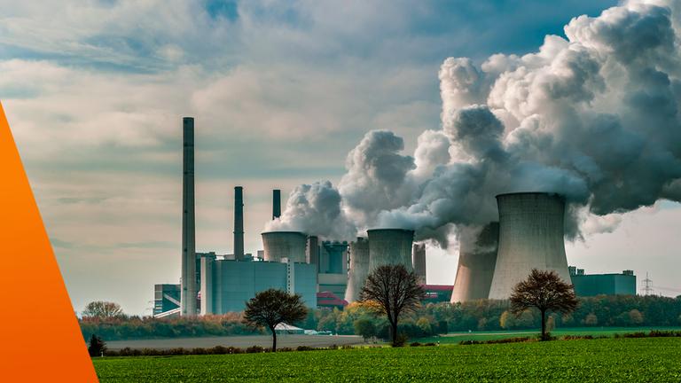 Zu sehen ist ein großes Kraftwerk mit mehreren Kühltürmen und Schornsteinen, aus denen dichte weiße Dampfwolken aufsteigen. Im Vordergrund liegen Felder und vereinzelte Bäume, darüber ein leicht bewölkter Himmel. Das Bild steht sinnbildlich für industrielle CO₂-Emissionen und die Herausforderungen des Emissionshandels.