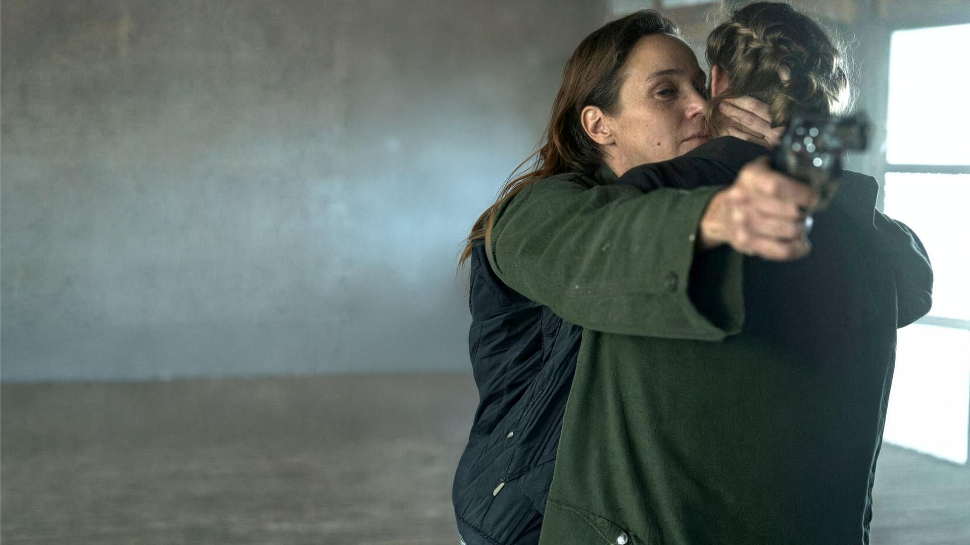 "Westwall - Kapitel VI": Ira (Jeanette Hain) hält Julia (Emma Bading) im Arm und zielt gleichzeitig mit einer Pistole auf die andere Seite des Raums.
