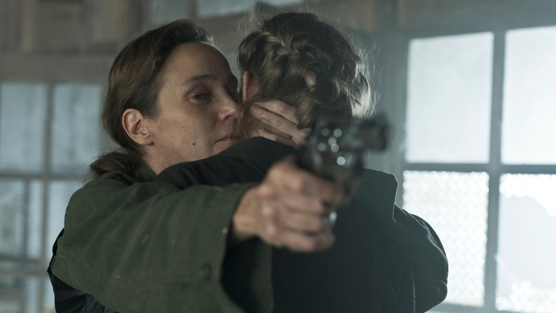 "Westwall - Kapitel VI": Ira (Jeanette Hain) hält Julia (Emma Bading) im Arm und zielt gleichzeitig mit einer Pistole auf die andere Seite des Raums.