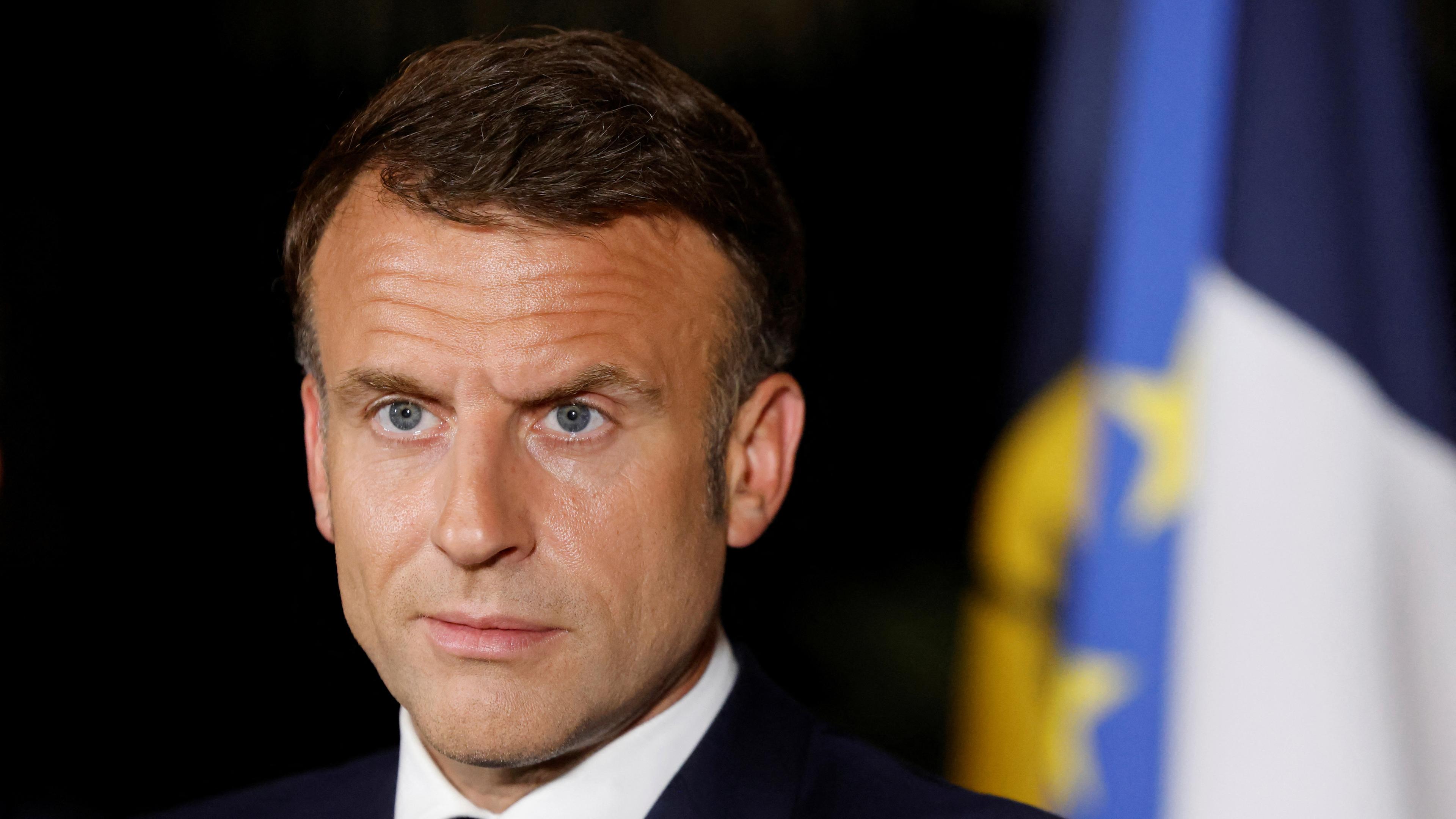 Der französische Präsident Macron steht vor einem schwarzen Hintergrund und schaut mit ernster Miene in die Ferne.