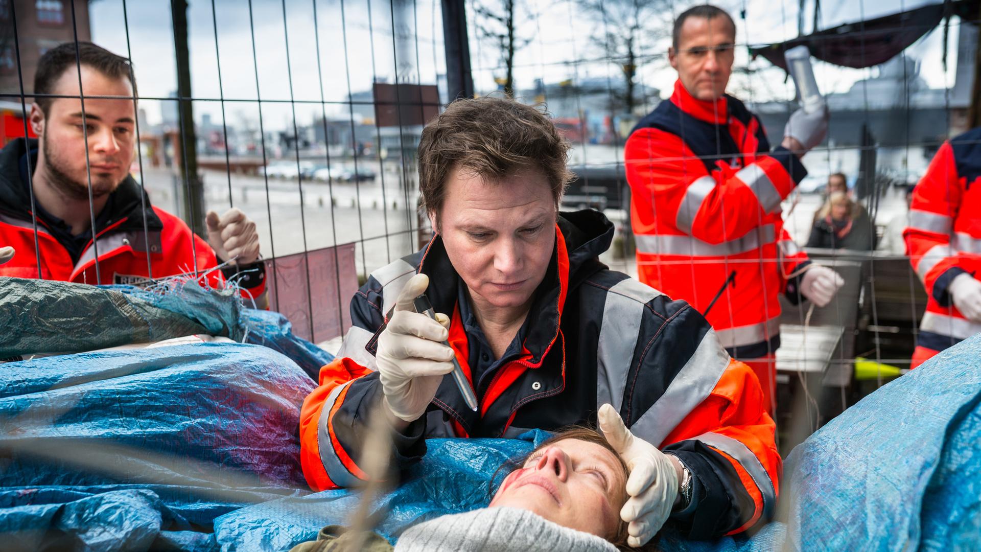"Notruf Hafenkante - Engel": Der Notarzt Dr. Haase (Fabian Harloff) steht hinter Nicole Wittich (Sabine Wolf), die auf blauen Planen auf dem Rücken liegt. Er hat seine linke Hand an ihrem Kopf und leuchtet mit der rechten mit einer Taschenlampe in ihre Augen.