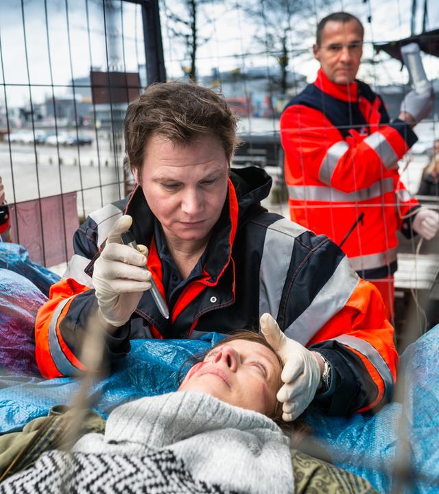 "Notruf Hafenkante - Engel": Der Notarzt Dr. Haase (Fabian Harloff) steht hinter Nicole Wittich (Sabine Wolf), die auf blauen Planen auf dem Rücken liegt. Er hat seine linke Hand an ihrem Kopf und leuchtet mit der rechten mit einer Taschenlampe in ihre Augen.