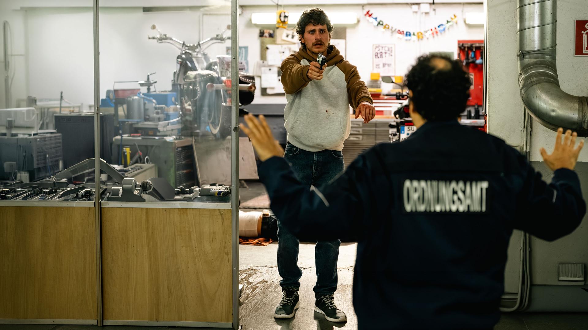"SOKO Köln - Engel": Maximilian Angerer (Ludwig Brix) steht in einer Werkstatt und richtet mit wütendem Blick eine Waffe auf Sinan Uzun (Ali Berber), der mit dem Rücken zur Kamera vor ihm steht. Sinan hat die Arme in kapitulierender Geste nach oben gehoben. Auf seiner Jacke ist verschwommen die Aufschrift "Ordnungsamt" zu lesen. Im Hintergrund sieht man Josephine Feiler (Hanni Lorenz) auf dem Boden der Werkstatt liegen. An der Wand ist eine "Happy Birthday"-Girlande befestigt.